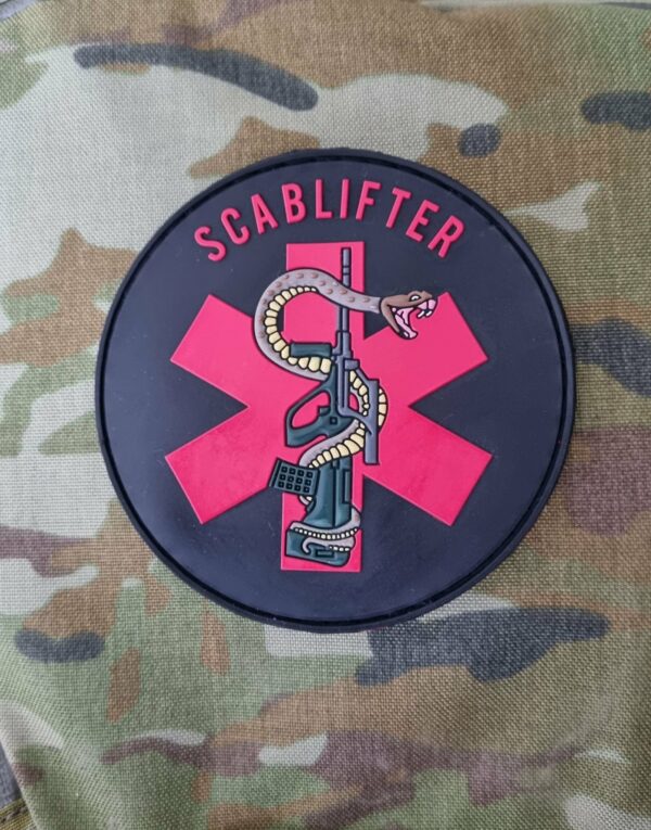 A Bored Scablifter Patch with a red cross and a serpent-entwined dagger, labeled "The Bored Scablifter Patch," displayed on a camouflage fabric background.