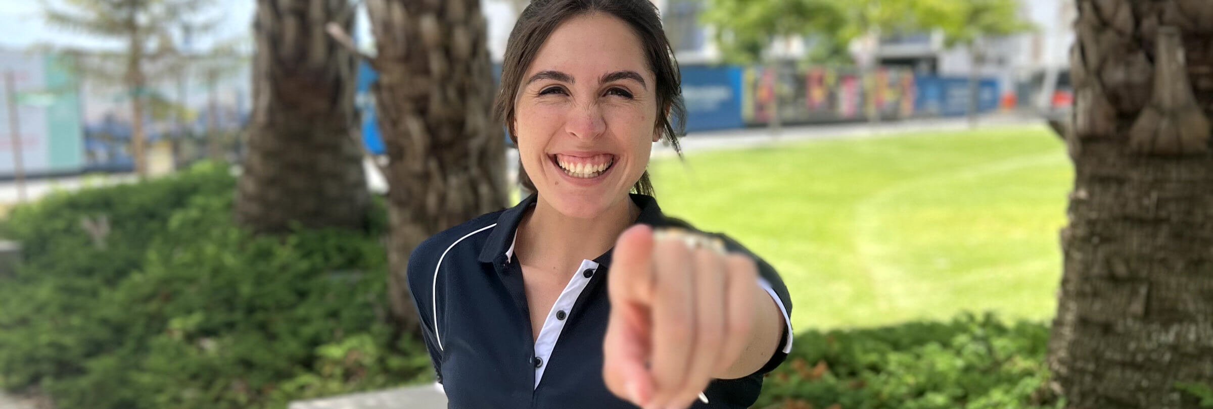A smiling woman pointing towards the camera with one hand, standing outdoors with trees and a blurred background.