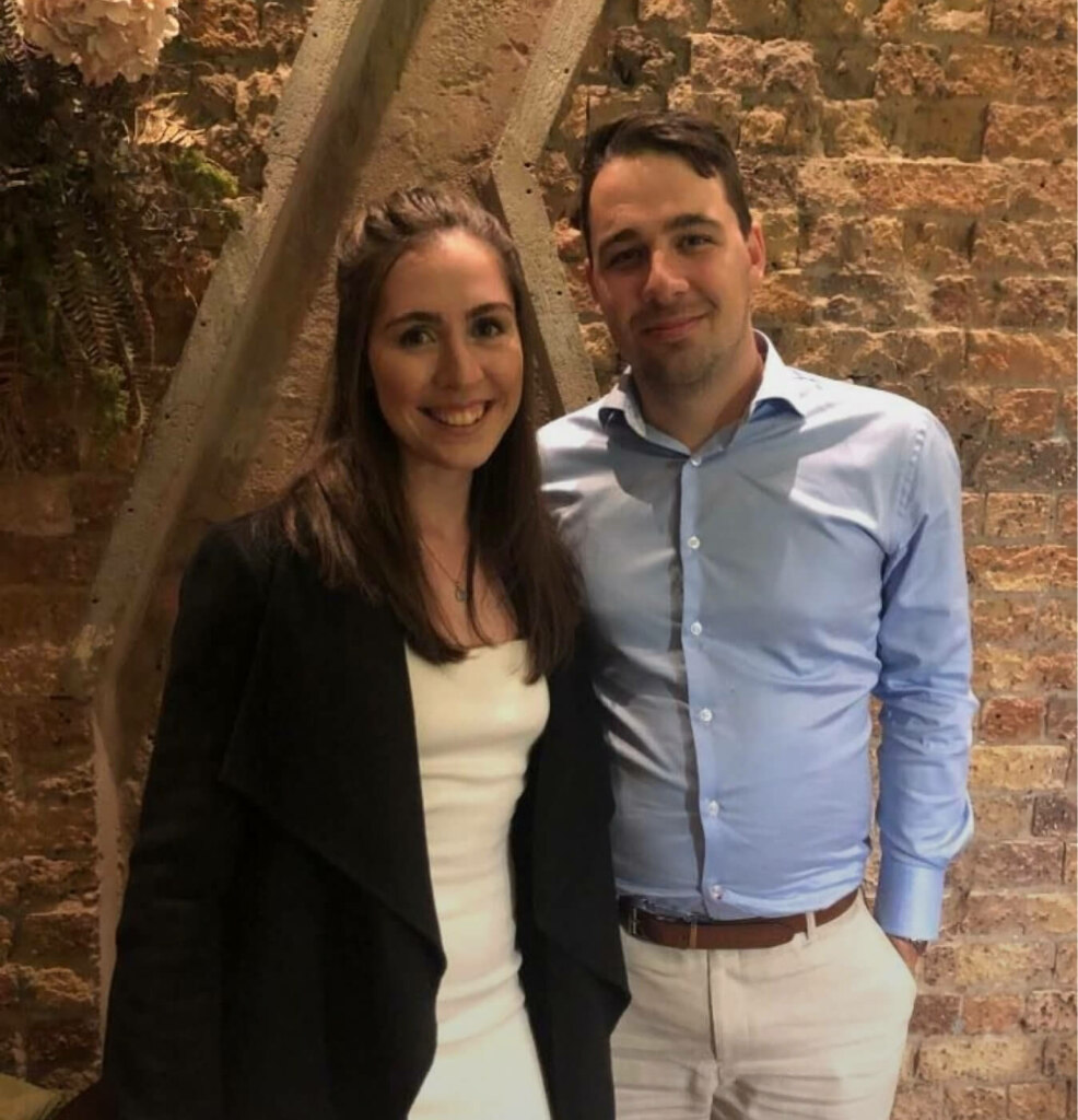 A man and woman posing together in front of a brick wall with a stair railing.